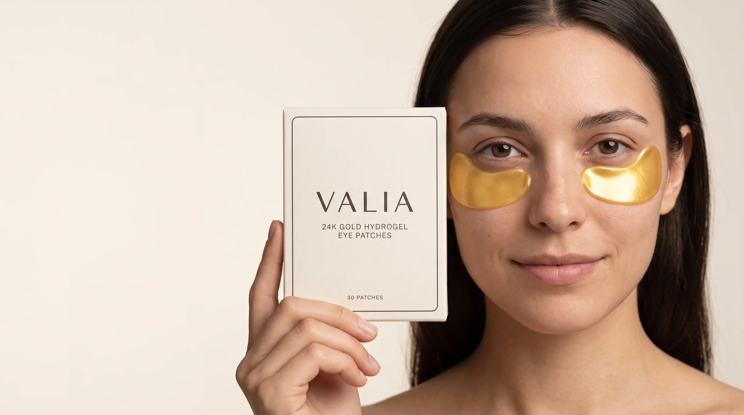 A woman holding the VALIA box beside her face, gold hydrogel crescents under each eye.