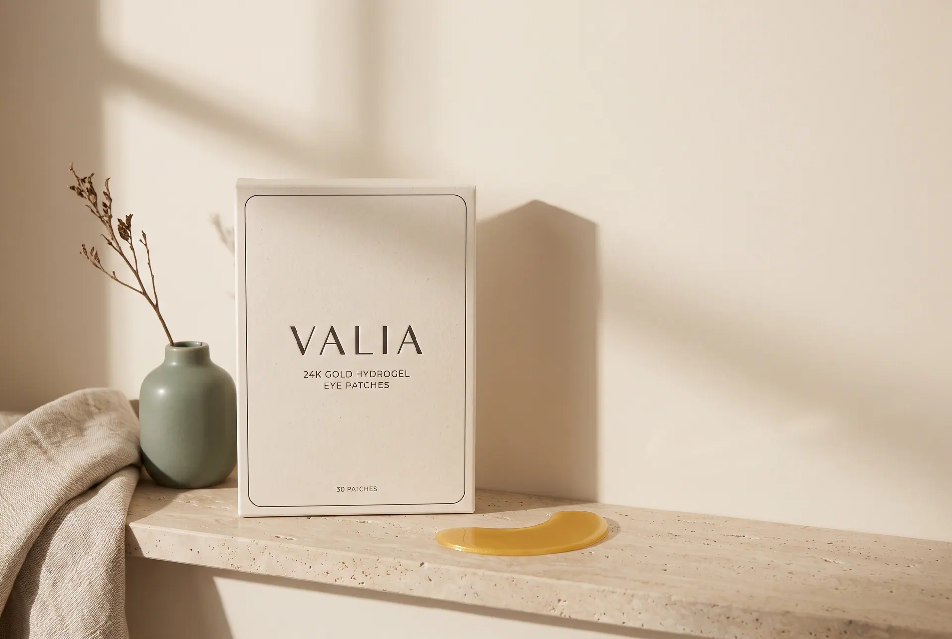 A travertine ledge holding the VALIA box, a sage ceramic vase, a folded linen towel and a single gold crescent.